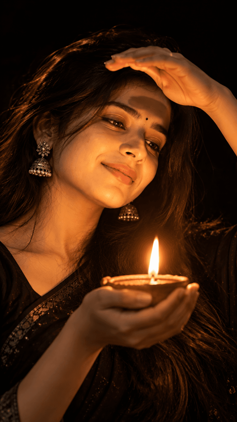 Woman in black traditional clothing holding a glowing clay diya, with silver jhumka earrings and a raised hand shielding her eyes.