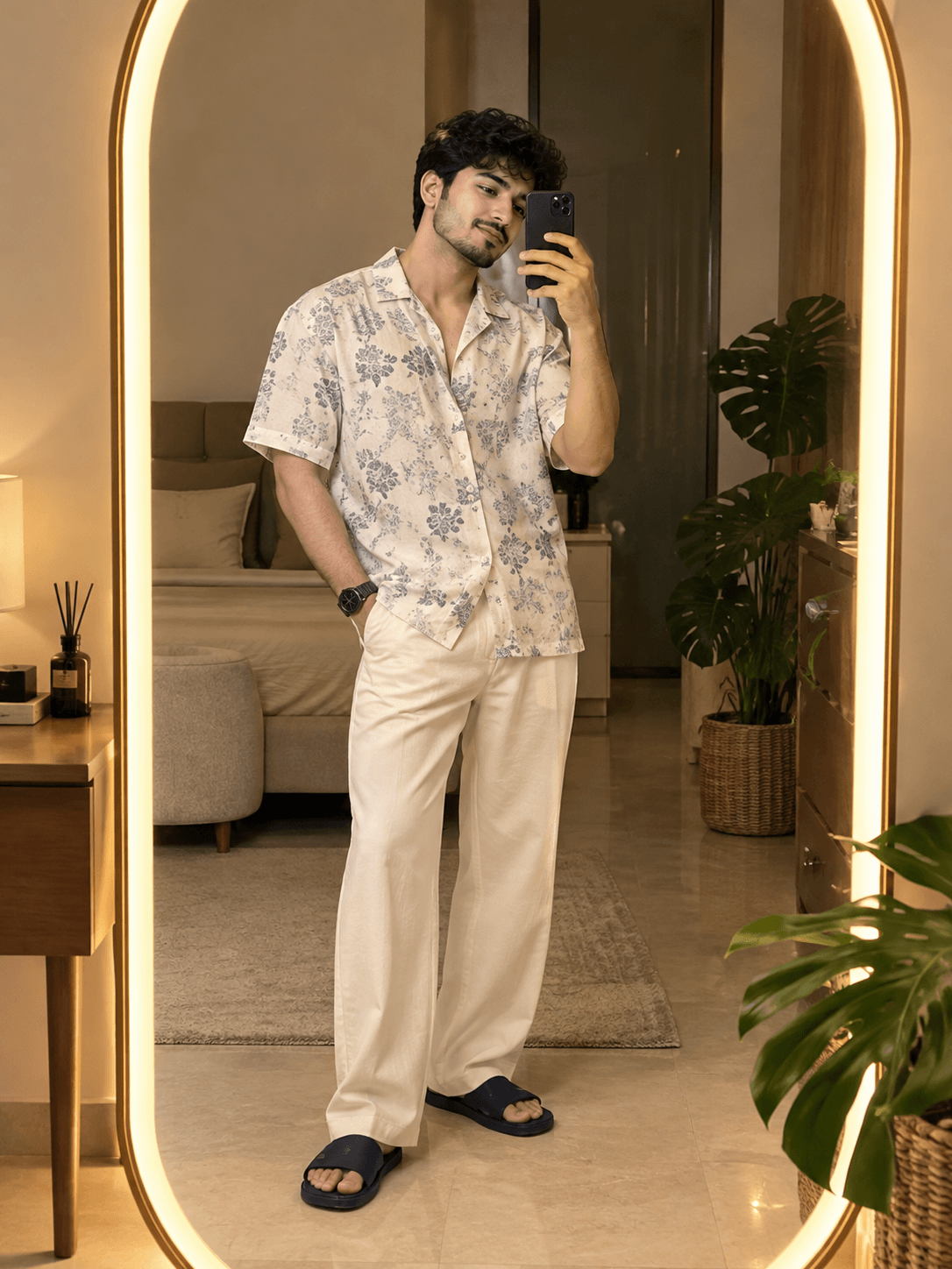 Young man in an off-white patterned shirt, cream trousers and navy slides taking a full-body selfie in an oval LED mirror.