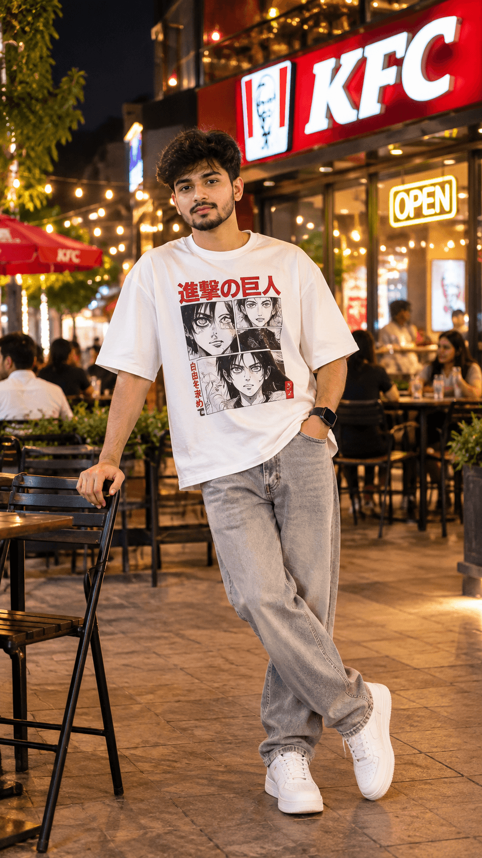 Young man in a white Japanese manga-print T-shirt, grey jeans and white sneakers posing beside a chair under KFC storefront lights at night.