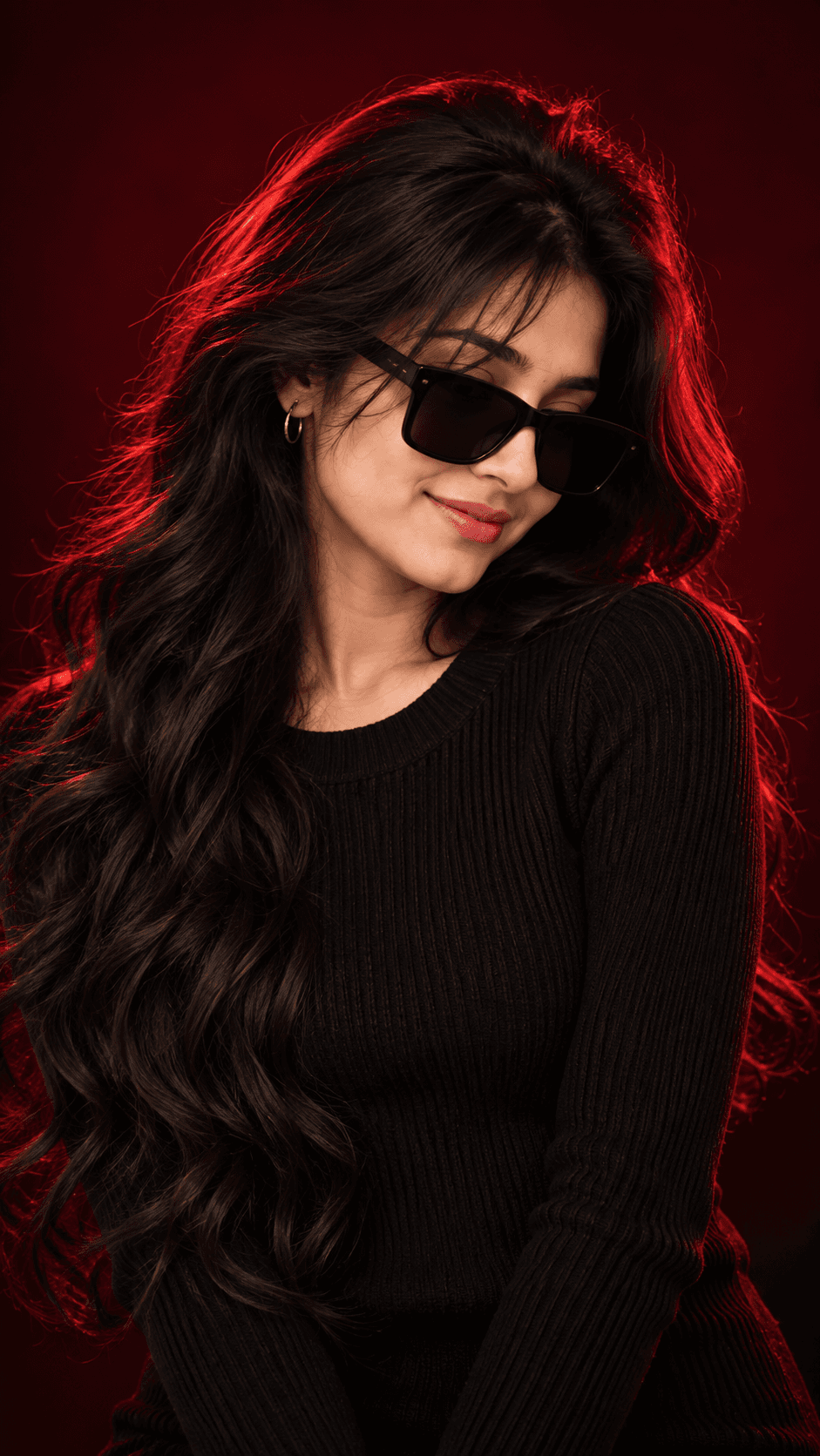 Woman with long wavy black hair, oversized black sunglasses and a ribbed sweater, lit by red rim lighting against a dark burgundy studio background.
