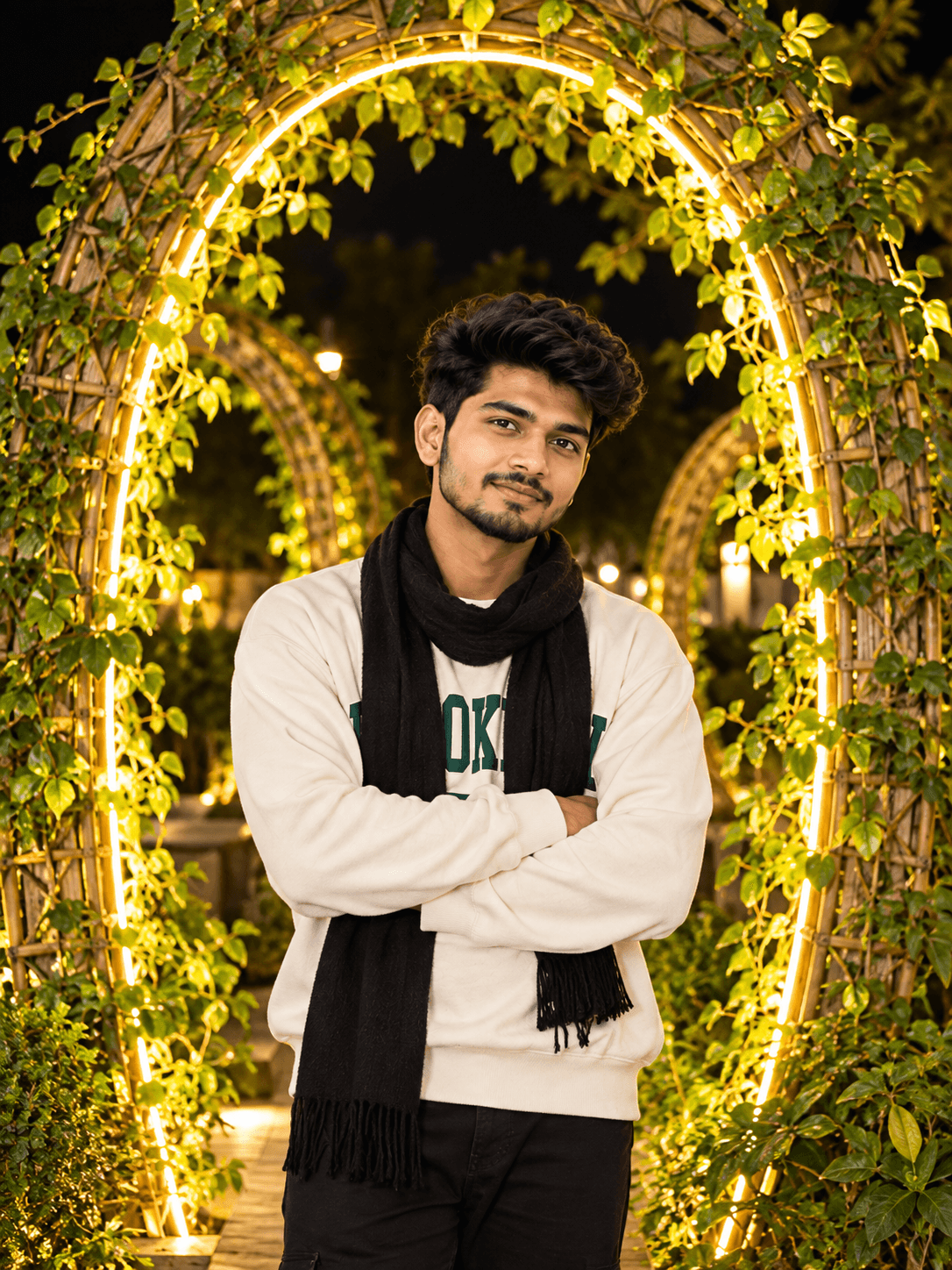 Young Indian man in an off-white sweatshirt and black scarf standing before a glowing garden arch covered with green vines at night.