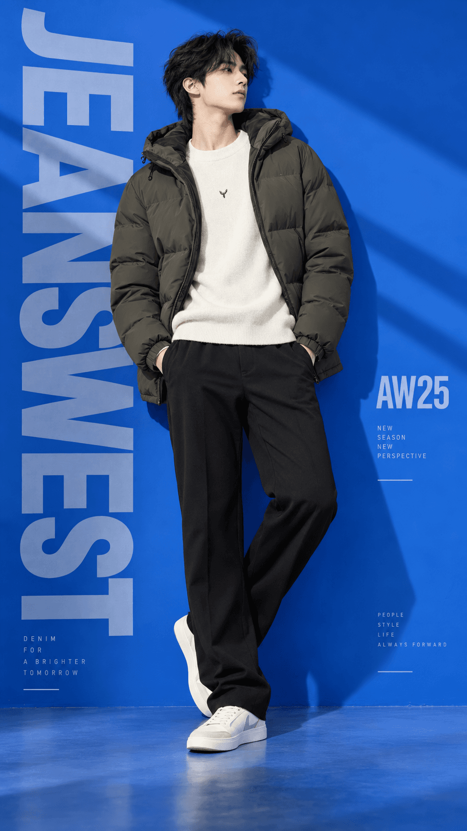 Young man in an olive puffer jacket, cream sweater and black trousers posing against a cobalt-blue JEANSWEST fashion studio backdrop.
